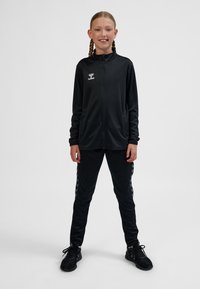 Svart sportjacka med hög krage och dragkedja, matchad med byxor som har ett kontrasterande chevronmönster. Bärs med svarta sneakers.