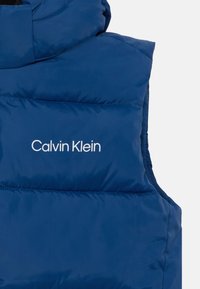 Modra puhovka z visoko ovratnikom, ki ima prešito teksturo in bel logotip "Calvin Klein" spredaj. Lahka material.