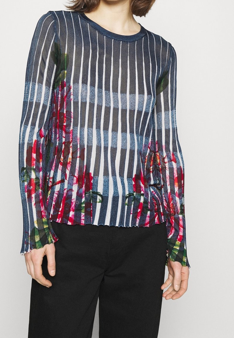 Femme portant un pull semi-transparent bleu marine à rayures blanches verticales et motifs floraux rouges, associé à un pantalon noir.