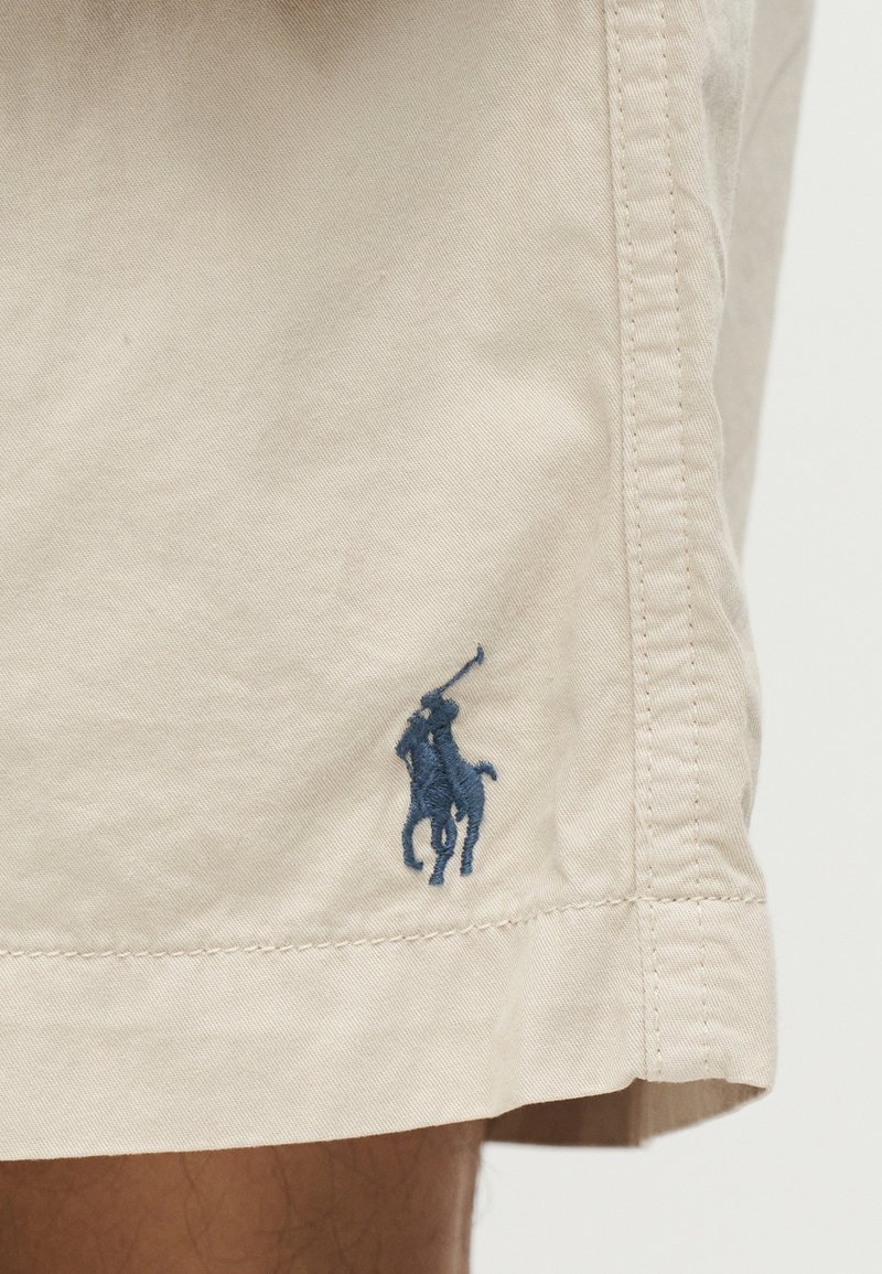 Ourlet de short beige avec un joueur de polo bleu brodé à cheval tenant un maillet, situé au-dessus de la jambe d'une personne.