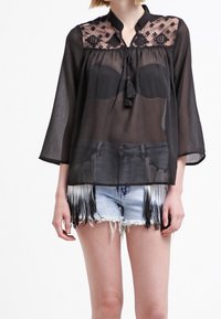 Zwarte transparante blouse met geborduurde kant yoke, mandarin kraag en 3/4 mouwen. Gecombineerd met versleten denim shorts met franjes.