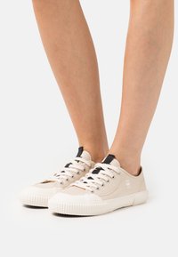 Beige canvas sneakers met witte rubberen zolen, ronde neus, zwarte hieltabs en contrasterende veters. Textuurstof en logo-accent.