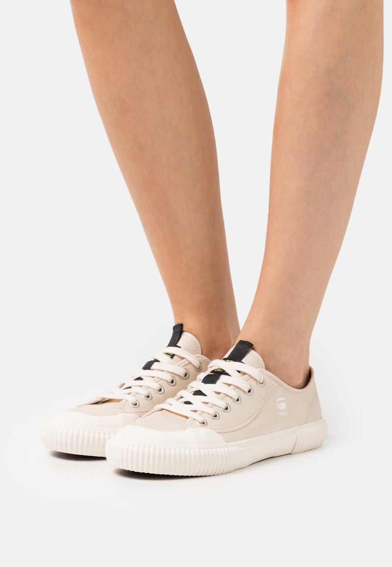 Beige canvas sneakers met witte rubberen zolen, ronde neus, zwarte hieltabs en contrasterende veters. Textuurstof en logo-accent.