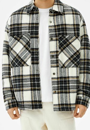 Mann trägt ein schwarz-weiß-beiges kariertes Hemd über einem schlichten weißen T-Shirt und beige Hose, Hände entspannt an den Seiten.