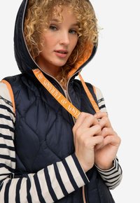 Gilet trapuntato blu navy con cappuccio, caratterizzato da una fodera arancione e un cordoncino. Una maglietta a righe a maniche lunghe è parzialmente visibile sotto il gilet.