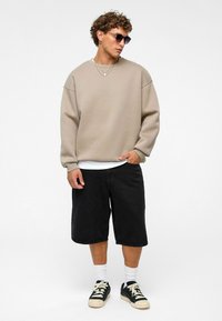 Beige oversized sweatshirt, zwarte denim shorts, zwarte sneakers met crèmekleurige accenten, gelaagde kettingen en zonnebril; casual streetwear look.