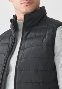 Black puffer vest with a high collar, quilted design, and front zipper. Worn over a light gray sweater, showcasing a smooth texture.
