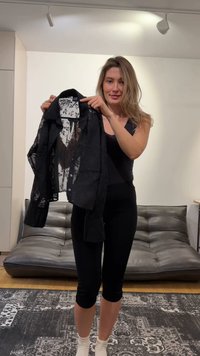 Vrouw in zwarte sportkleding die een zwart doorschijnend shirt met een bloemenprint aan de binnenkant vasthoudt, staand in een moderne woonkamer met een grijze leren bank.