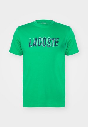 Camiseta de algodón verde con mangas cortas y cuello redondo, que presenta un destacado logo bordado en azul de "LACOSTE" en el pecho.