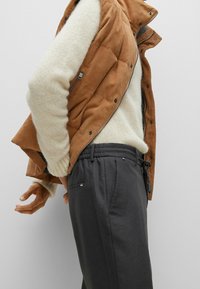 Chaleco de ante marrón sobre un suéter crema, combinado con pantalones de cintura elástica gris oscuro que cuentan con un bolsillo y cordón. Tejidos texturizados visibles.