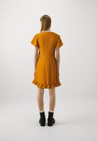 Robe jaune moutarde avec un motif de diamants texturé, manches courtes à volants et ourlet froncé. Portée avec des bottines noires et des chaussettes blanches.