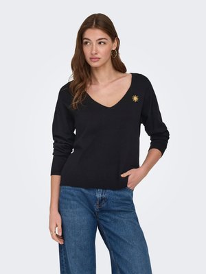 Mujer que lleva un suéter negro con cuello en V y un pequeño emblema amarillo de sol en el pecho, y vaqueros azules, de pie con una mano en el bolsillo.