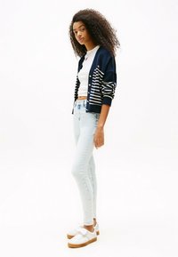 Cárdigan azul marino con rayas blancas, combinado con una camisa blanca ajustada, jeans ajustados de color azul claro y zapatillas blancas con suela marrón.