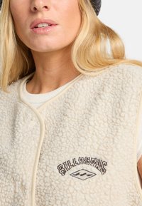 Cream fleece vest with a textured surface, embroidered logo in black and beige accents, and a contrasting collar design.