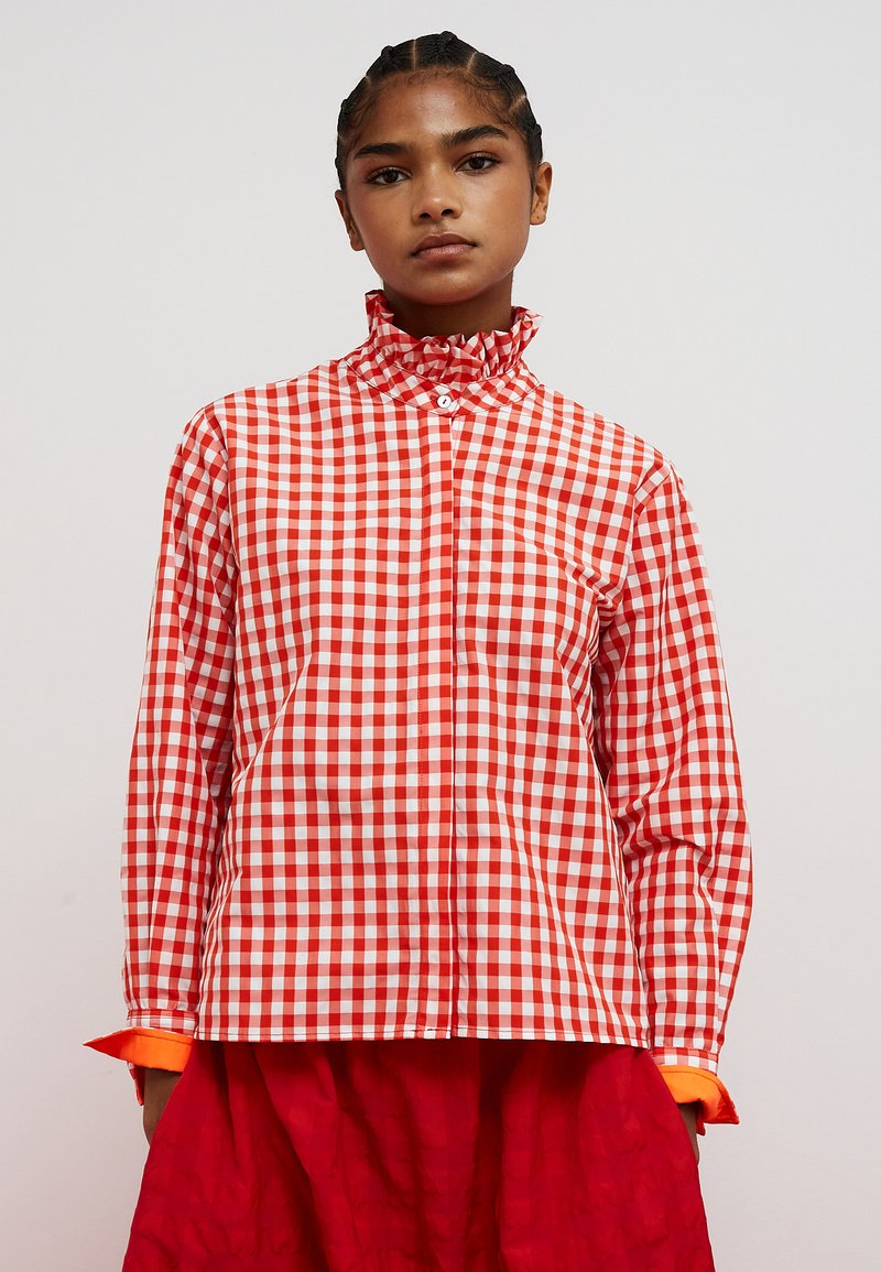 Red and white gingham blouse with a high ruffled collar, long sleeves, and orange cuff accents. The fabric is smooth with a glossy finish.