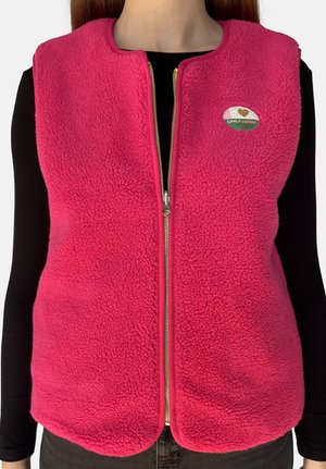 Woman wearing a bright pink zip-up fleece vest over a black long-sleeve top, with a small oval patch on the chest.