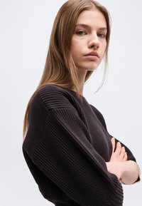 Νεαρή γυναίκα με μακριά ανοιχτό καστανά μαλλιά, φοράει μαύρο πουλόβερ με υφή, με σταυρωμένα τα χέρια, κοιτώντας με αυτοπεποίθηση την κάμερα.