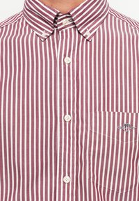 Bordeauxrood en witte verticaal gestreepte overhemd met knoopsluiting en een button-down kraag, en een borstzak geborduurd met het "GANT" logo.