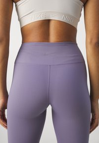 Femme portant un soutien-gorge de sport blanc Nike et un legging lavande taille haute Nike, vue de dos et de la taille vers le bas.