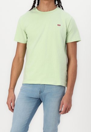 Henkilö, joka on pukeutunut vaaleanvihreään Levi's-t-paitaan ja vaalean sinisiin farkkuihin, seisoo valkoista taustaa vasten rentoutunein käsin.