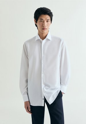 Joven con el pelo negro que lleva una camisa blanca de manga larga abotonada y pantalones negros, de pie contra un fondo blanco liso.