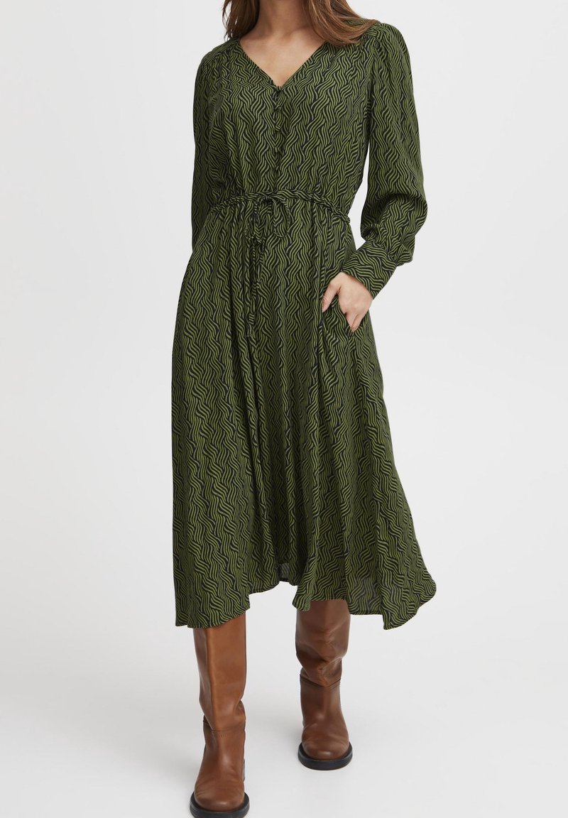 Femme portant une robe midi verte à motifs avec manches longues et ceinture à la taille, debout avec une main dans la poche et des bottes marron montant jusqu'aux genoux.