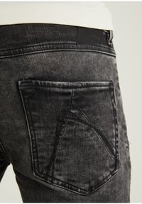 Lähikuva mustista haalistuneista farkuista, joissa korostuu takatasku ja ompeleet. Yläosana valkoinen paita, joka on taitettu farkkuihin.