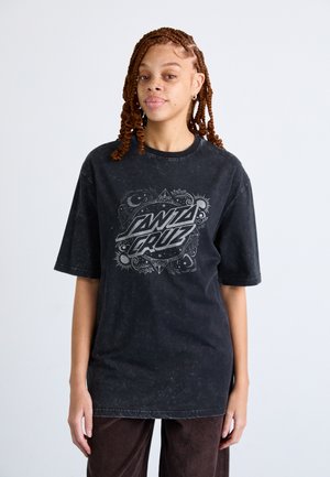 T-shirt oversize noir avec une texture usée, présentant un design graphique central en texte gris. Associé à un pantalon en velours côtelé marron.