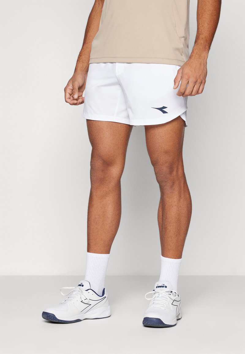 Weiße Sportshorts mit seitlichen Schlitzen, versehen mit einem blauen Logo. Kombiniert mit weißen Schuhen und weißen Kniestrümpfen. Darüber ein beiges T-Shirt.