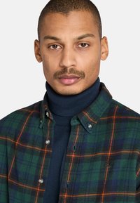 Groen geruite flanellen shirt met oranje en blauwe accenten, knoopsluiting bij de kraag, over een marineblauwe coltrui. De stof lijkt zacht met een gestructureerde pasvorm.