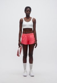 Γυναίκα στέκεται κρατώντας ροζ λουρί αντίστασης, φορώντας λευκό αθλητικό σουτιέν, κοραλλί αθλητικές σορτς, λευκές κάλτσες και λευκά αθλητικά παπούτσια σε απλό φόντο.