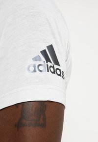 Vit t-shirt med kort ärm och en svart Adidas-logotyp tryckt på vänster axel. Tyget verkar mjukt med en slät textur.