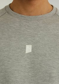 Grauer Sweatshirt mit strukturierter Oberfläche, ausgestattet mit einem kleinen weißen geometrischen Logo im Brustbereich und einem gerippten Halsausschnitt.