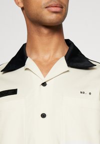 Camicia crema a maniche corte con colletto nero, taschino frontale e ricamo "NO. 0", caratterizzata da due bottoni neri e un tessuto testurizzato.