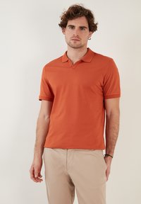 Oranžové polo tričko s límečkem, krátkým rukávem a hladkou texturou. Nosené s světle béžovými kalhotami, které mají volný střih a jednoduchý design.