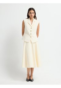 Elfenben ærmeløs vest med gyldne knapper, parret med nederdel i matchende midi længde. Glat stof, struktureret form, minimalistik design. Sorte sko.