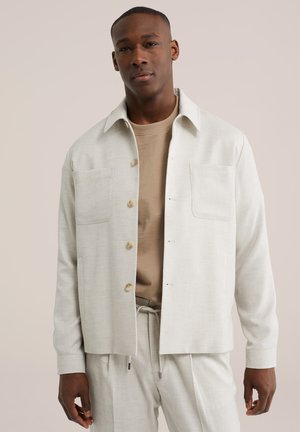 Mand iført beige jakke med knapper over tan skjorte og matchende beige bukser, stående foran en ensfarvet lys baggrund.