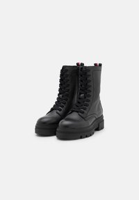 Botas de combate de cuero negro con un acabado texturizado, diseño de cordones, suela de goma gruesa y una lengüeta contrastante que presenta detalles en rojo y blanco.