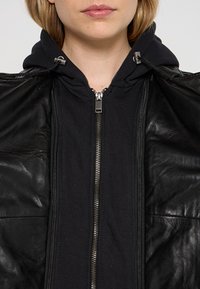 Černá kožená bunda s vysokým límcem, pod kterou je černá mikina na zip. Obsahuje kovové zipy a má hladkou strukturu.