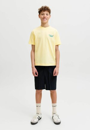 Tínedžer chlapec se usmívá, má na sobě žluté tričko s nápisem, černé krátké kalhoty s proužky, krémové ponožky s logy a bílé tenisky, stojí před jednoduchým pozadím.