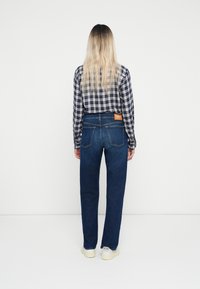 Denim jeans i mørkeblå, rette ben. Kombinert med en svart og hvit rutete skjorte, som har en brun lær etikett på baksiden.
