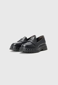 Schwarze Lederschlupfschuhe mit einem erhöhten Vorderteil, klobiger Sohle und metallischen Nietenakzenten am Boden. Glatte Textur und rundes Zehendesign.