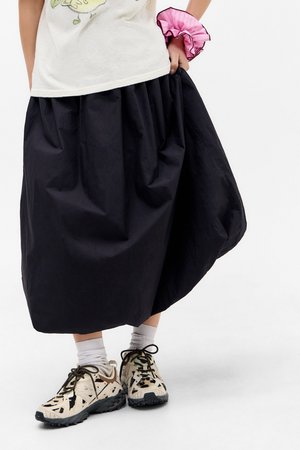 Gonna nera fino al ginocchio con un design arricciato, realizzata in materiale leggero. Indossata con sneakers a fantasia e calzini bianchi alla caviglia.