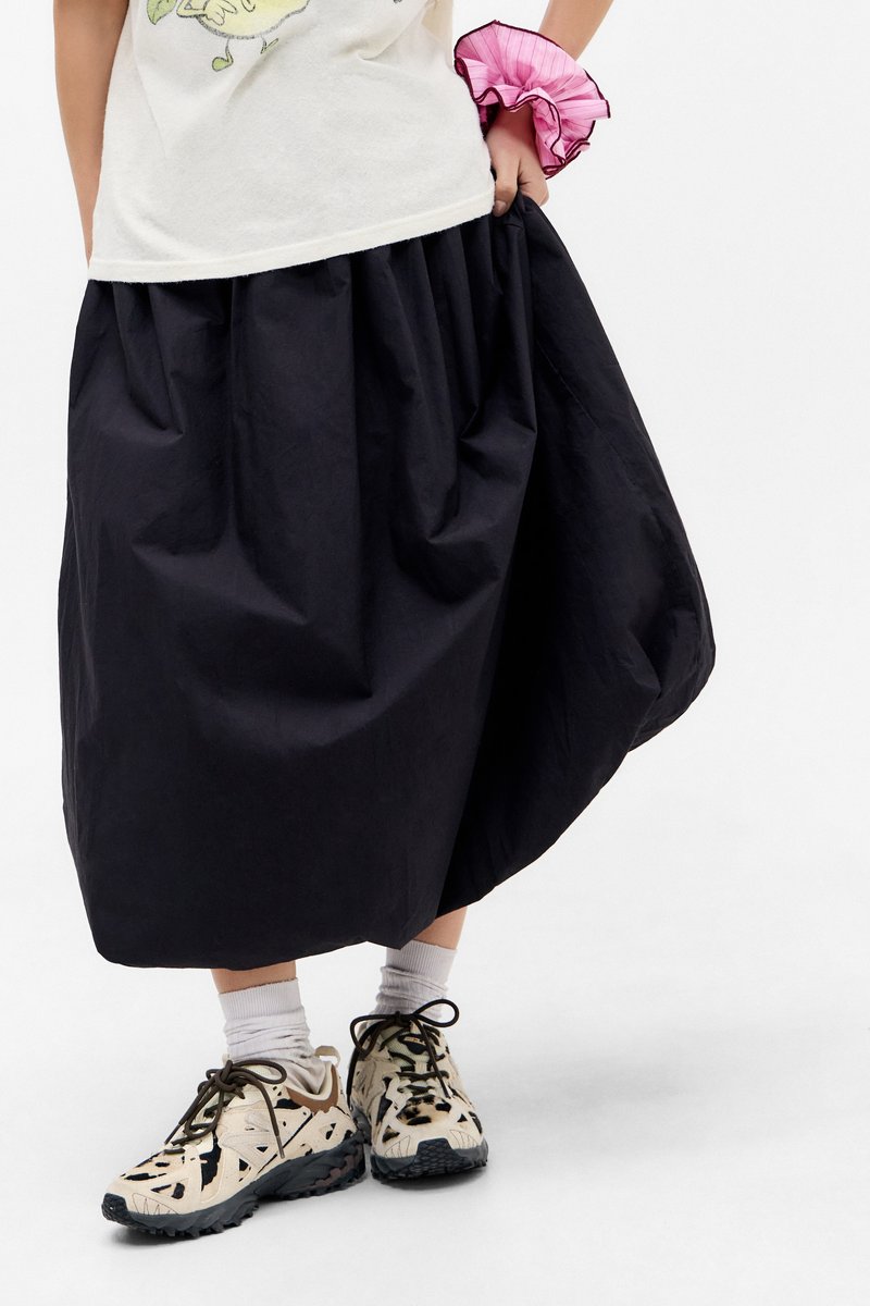Gonna nera fino al ginocchio con un design arricciato, realizzata in materiale leggero. Indossata con sneakers a fantasia e calzini bianchi alla caviglia.