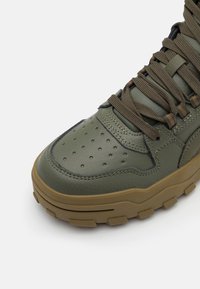 Baskets en cuir vert olive avec un design perforé, semelle en caoutchouc marron épaisse et lacets assortis. Présente des détails cousus et une texture robuste.