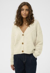 Kremasti pleteni kardigan s V-izrezom, prednjim gumbima i dekorativnim uzorcima spirala. Rebrasti manžeti i rub. Uparen s tamnoplavim trapericama.