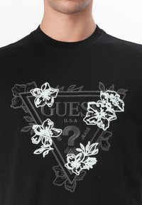 Black t-shirt featuring a triangular graphic with the word "GUESS" and detailed white floral embroidery around it.