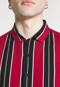 Zbliżenie na dolną część twarzy i szyję osoby noszącej koszulę w pionowe paski w kolorach czerwonym, czarnym i białym zapinaną na guziki oraz srebrne kolczyki koła.