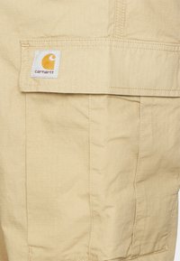 Beige Cargo-Shorts mit strukturiertem Gewebe, mehreren Taschen und einem gestickten Carhartt-Logoetikett auf der rechten Seite.