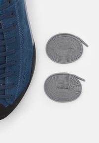 Niebieskie buty wspinaczkowe z zamszowej skóry z ciemną gumową podeszwą i szarymi sznurowadłami, z bocznym brandingiem i gładkim wykończeniem. W zestawie znajdują się dwa dodatkowe szare sznurowadła.